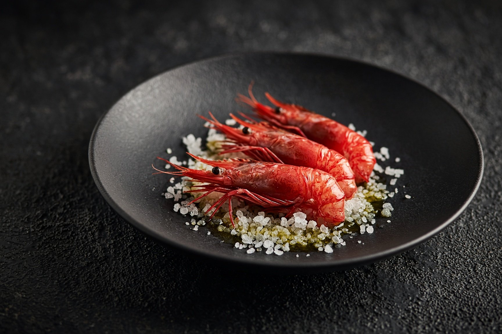 Three raw red prawns, heads on, on coarse sea salt and oil on a matt black plate — illustrative AI-generated demo image.