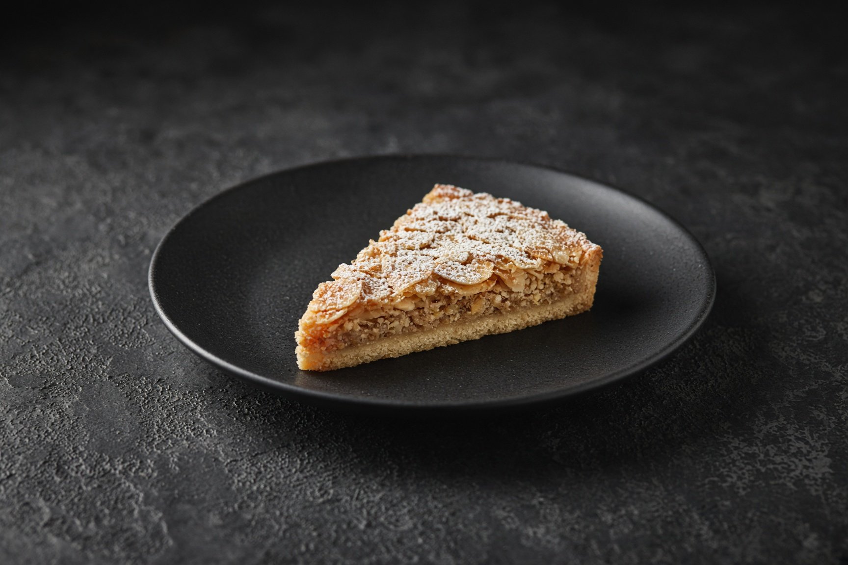 A wedge of almond tart, flaked almonds and icing sugar, on a matt black plate — illustrative AI-generated demo image.