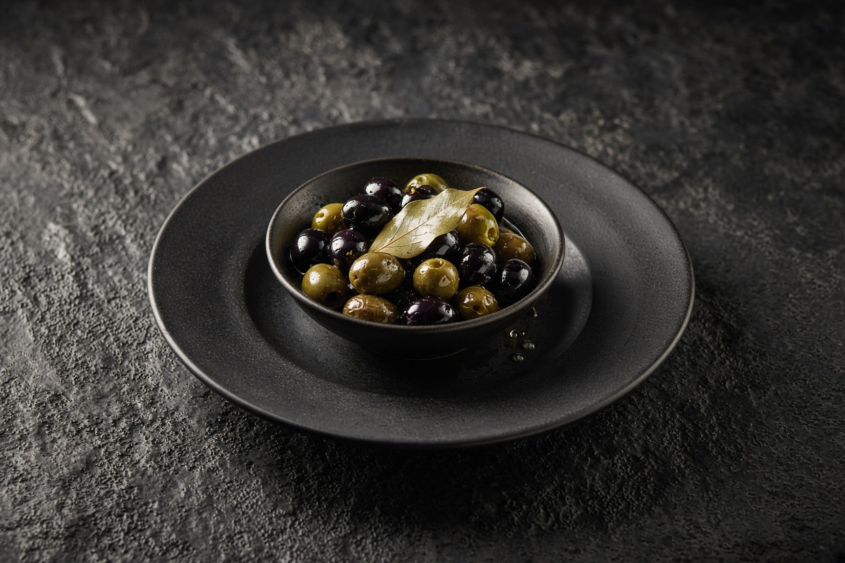 A small dark bowl of green and black olives in oil with a bay leaf, on a matt black plate — illustrative AI-generated demo image.