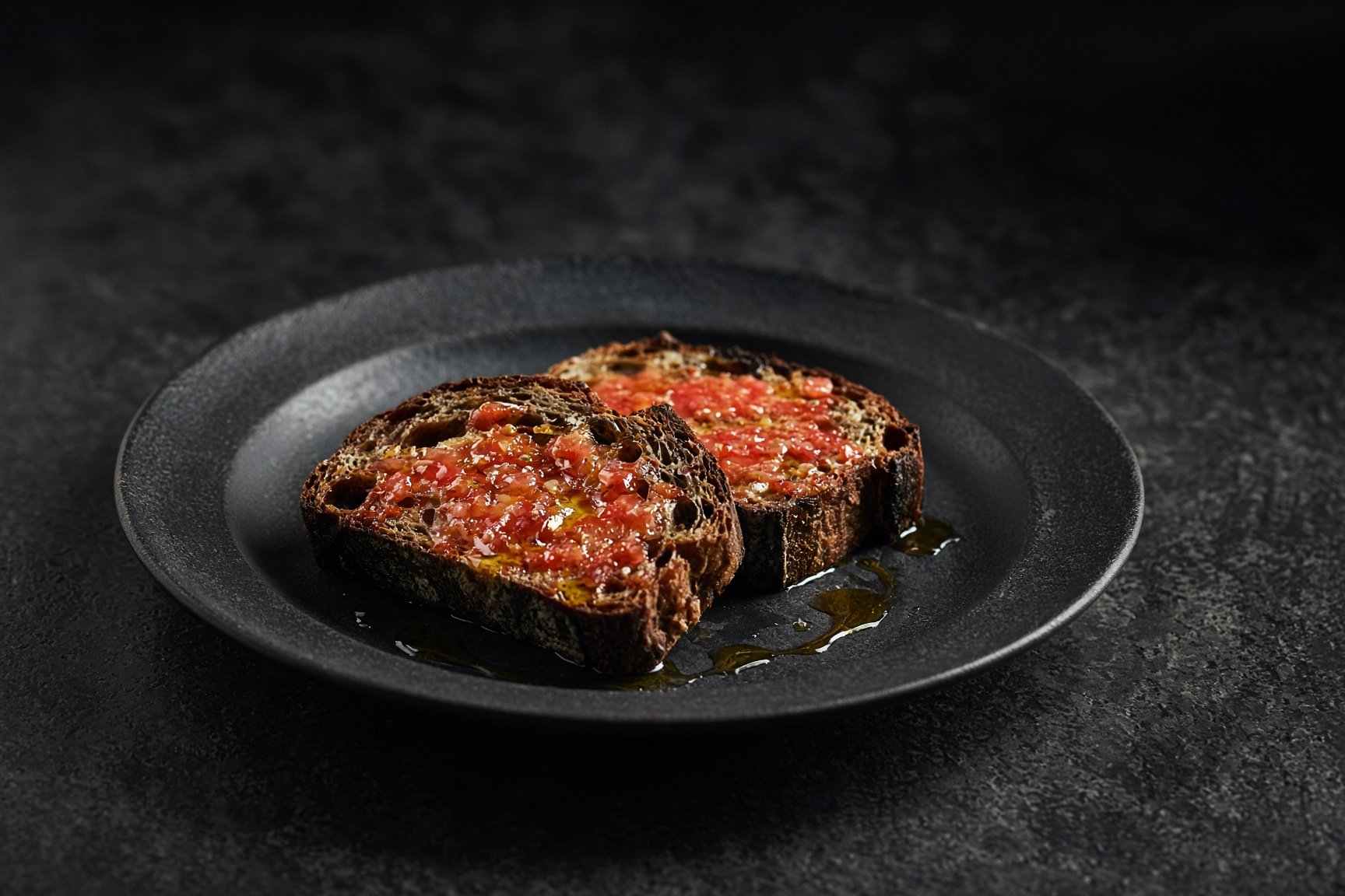 Two slices of dark country bread rubbed with tomato, olive oil pooling on a matt black plate — illustrative AI-generated demo image.